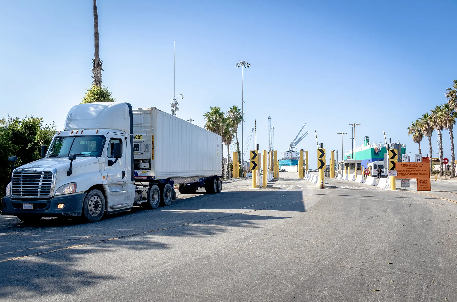 A semi-truck with a white trailer approaches a security checkpoint with barriers and yellow signs, ensuring compliance with port regulations. Palm trees and port cranes are visible in the background under a clear blue sky.