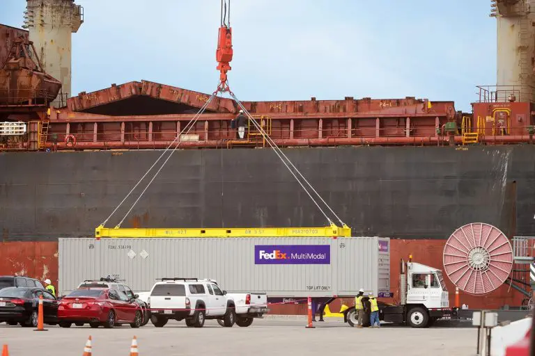 A large cargo container, marked with the FedEx logo, is being carefully lifted by a crane onto a container vessel at the Port of Hueneme. Workers, vehicles, and equipment bustle on the dock below.