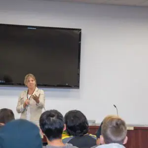 A person speaks to an audience in a conference room with a large blank screen and microphones on a table. The California state flag is visible on the right side, and Kristin Decas is among the attentive listeners.