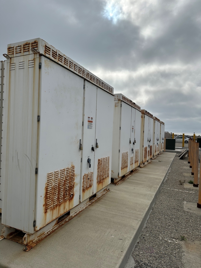 A row of large white metal utility cabinets with visible rust stains stands on a concrete walkway under a cloudy sky.