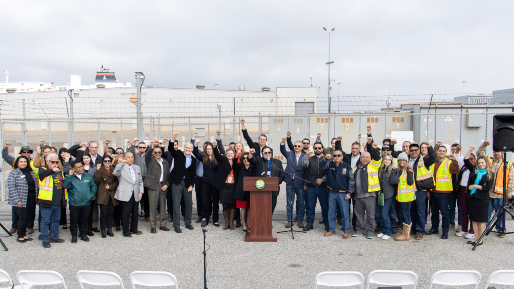 A large group of people, some in safety vests, stand behind a podium outdoors, raising their fists in the air near a fenced industrial area.