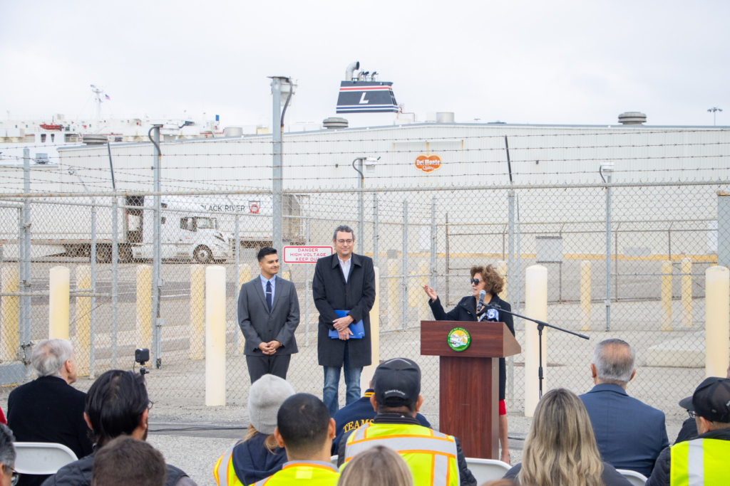A woman speaks at a podium in front of a fenced industrial area, with two men standing beside her and an audience seated in front.
