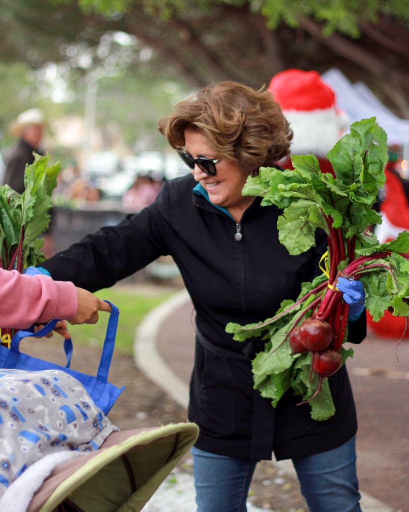 A woman wearing sunglasses hands a bunch of beets to another person outdoors, with trees and people visible in the background.
