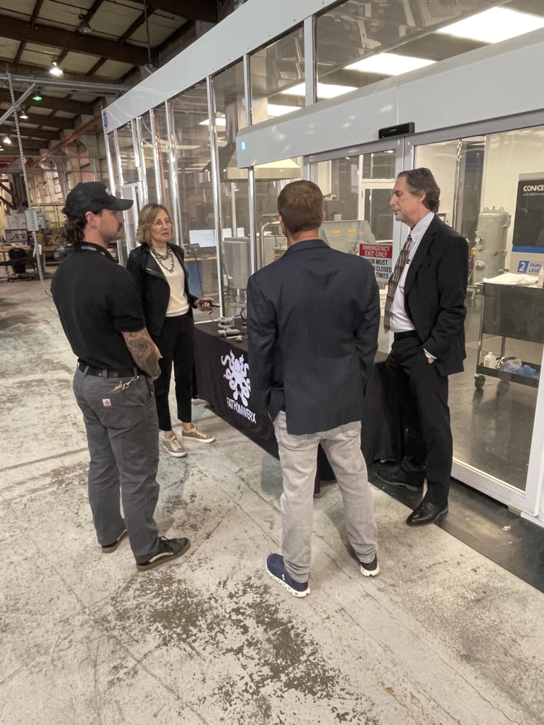 Four people stand and converse in an industrial facility near glass-walled enclosures, with machinery visible inside the room.