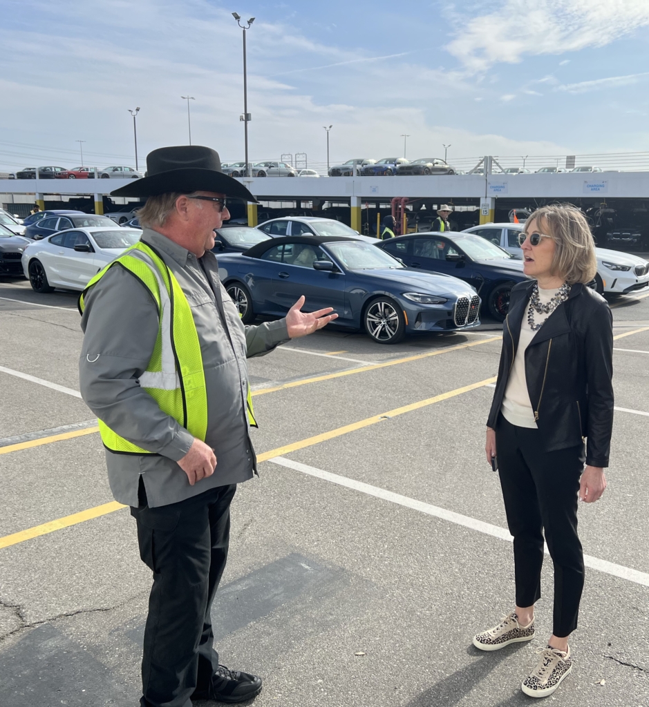A man in a safety vest and hat speaks to a woman in sunglasses and a black jacket in a parking lot with several parked cars in the background.