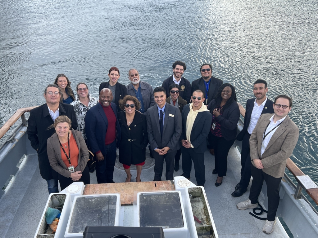 A group of fifteen people, dressed in business and casual attire, stand and pose for a photo on the deck of a boat with water visible in the background.