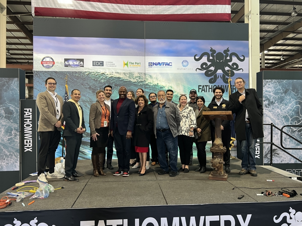 A group of people poses on a stage in front of a FATHOMWERX backdrop at an indoor event, with logos and an American flag visible in the background.