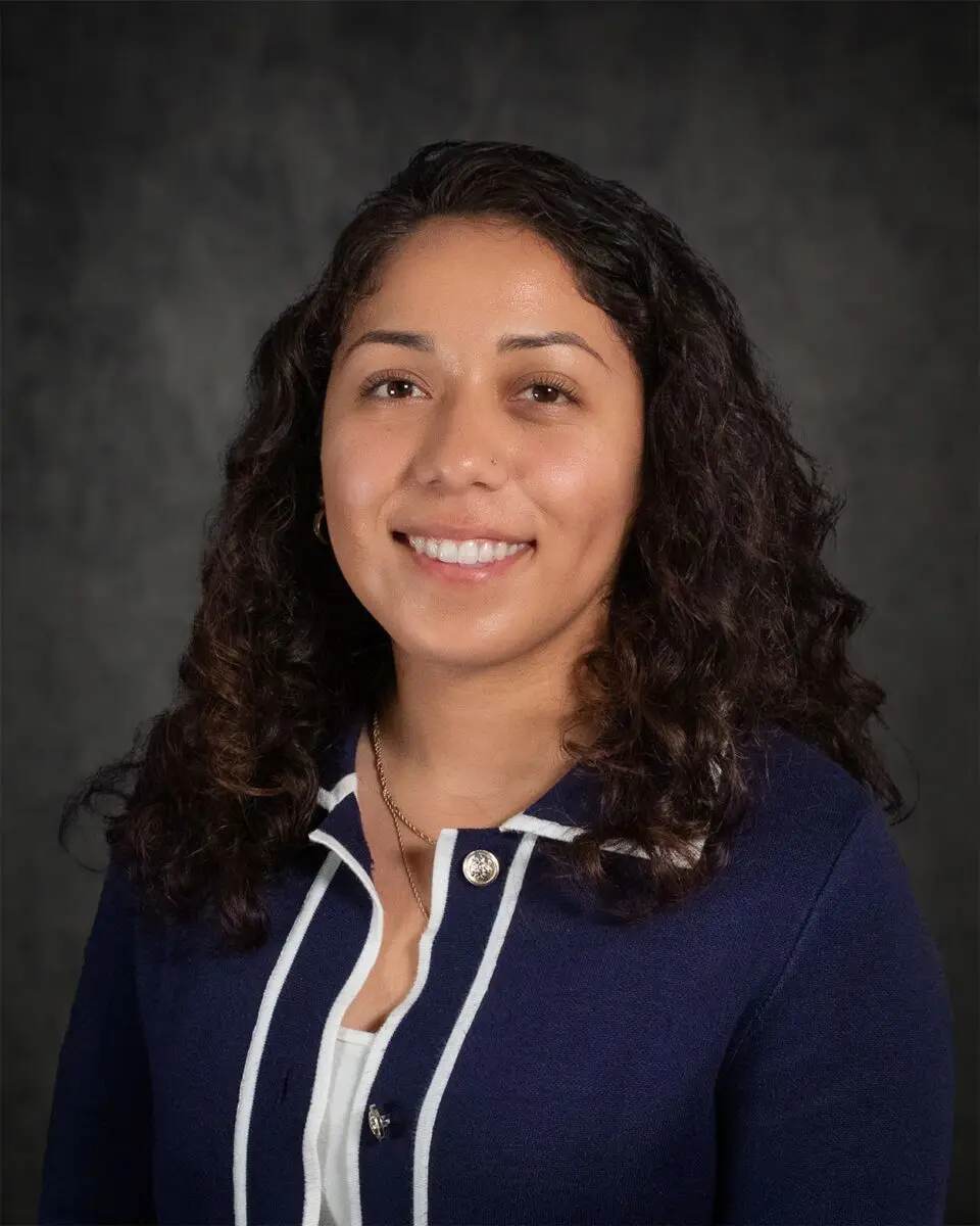 A woman with curly dark hair, smiling, dressed in a navy blue jacket with white trim, stands against a dark background reminiscent of Jacobo's signature style.