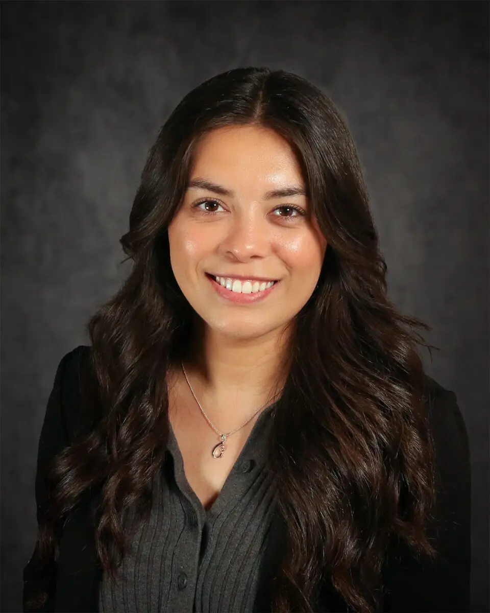 Eliana Gonzales, a woman with long brown hair, smiles at the camera, wearing a dark gray button-up top and a black jacket, with a pendant necklace on a dark background.