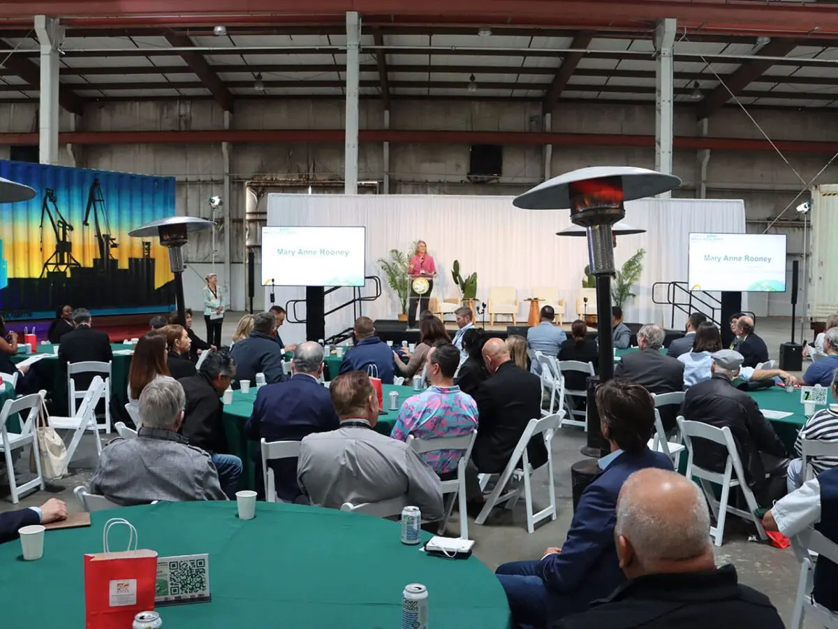 A speaker addresses an audience seated at round tables in a large indoor venue, with two presentation screens and industrial heaters visible, during the announcement of a $3.935 Million State Grant for Port of Hueneme as Governor ramps up the California Port Data Partnership Program.