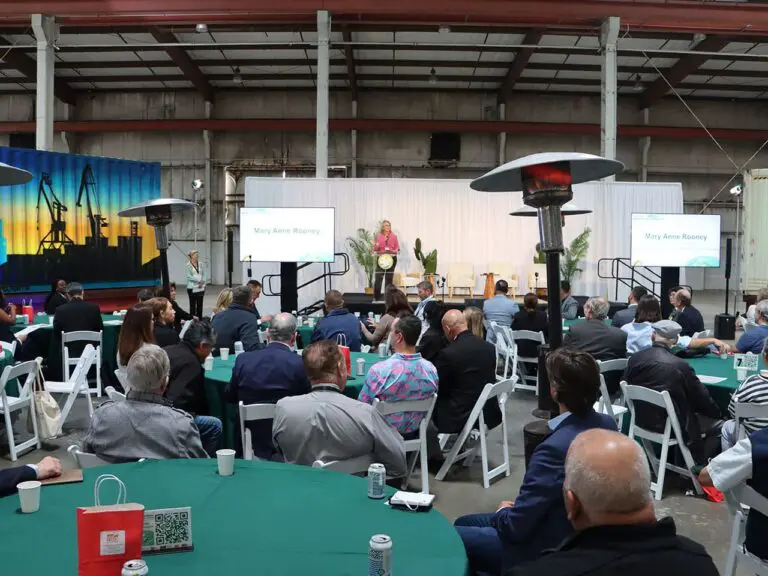 A speaker addresses an audience seated at round tables in a large indoor venue, with two presentation screens and industrial heaters visible, during the announcement of a $3.935 Million State Grant for Port of Hueneme as Governor ramps up the California Port Data Partnership Program.