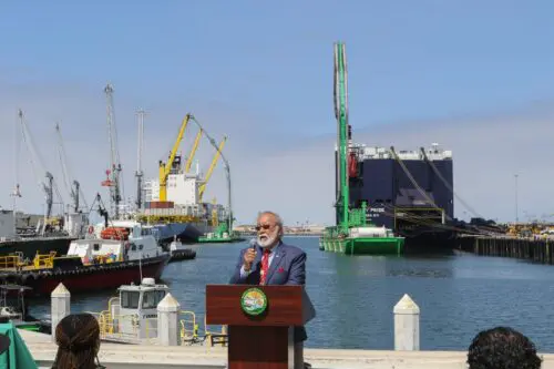 A man in a suit speaks at a podium by a harbor with cranes and cargo ships, announcing a $3.935 Million State Grant for Port of Hueneme as Governor ramps up the California Port Data Partnership Program.