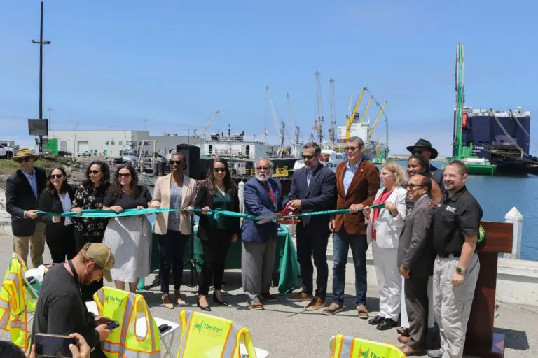 A group of people stand in a line cutting a ceremonial ribbon at the Port of Hueneme, celebrating a $3.935 Million State Grant as Governor ramps up the California Port Data Partnership Program, with cranes and cargo ships in the background.