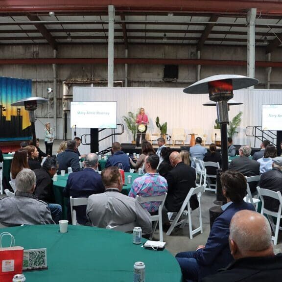 A speaker addresses an audience seated at round tables in a large indoor venue, with two presentation screens and industrial heaters visible, during the announcement of a $3.935 Million State Grant for Port of Hueneme as Governor ramps up the California Port Data Partnership Program.